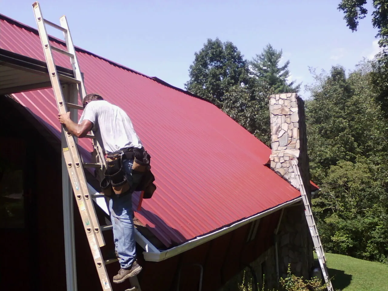 Roofer on a ladder beside a red metal roof and stone chimney — Soffit & Fascia Repair by Roofing Brothers in Pearlington